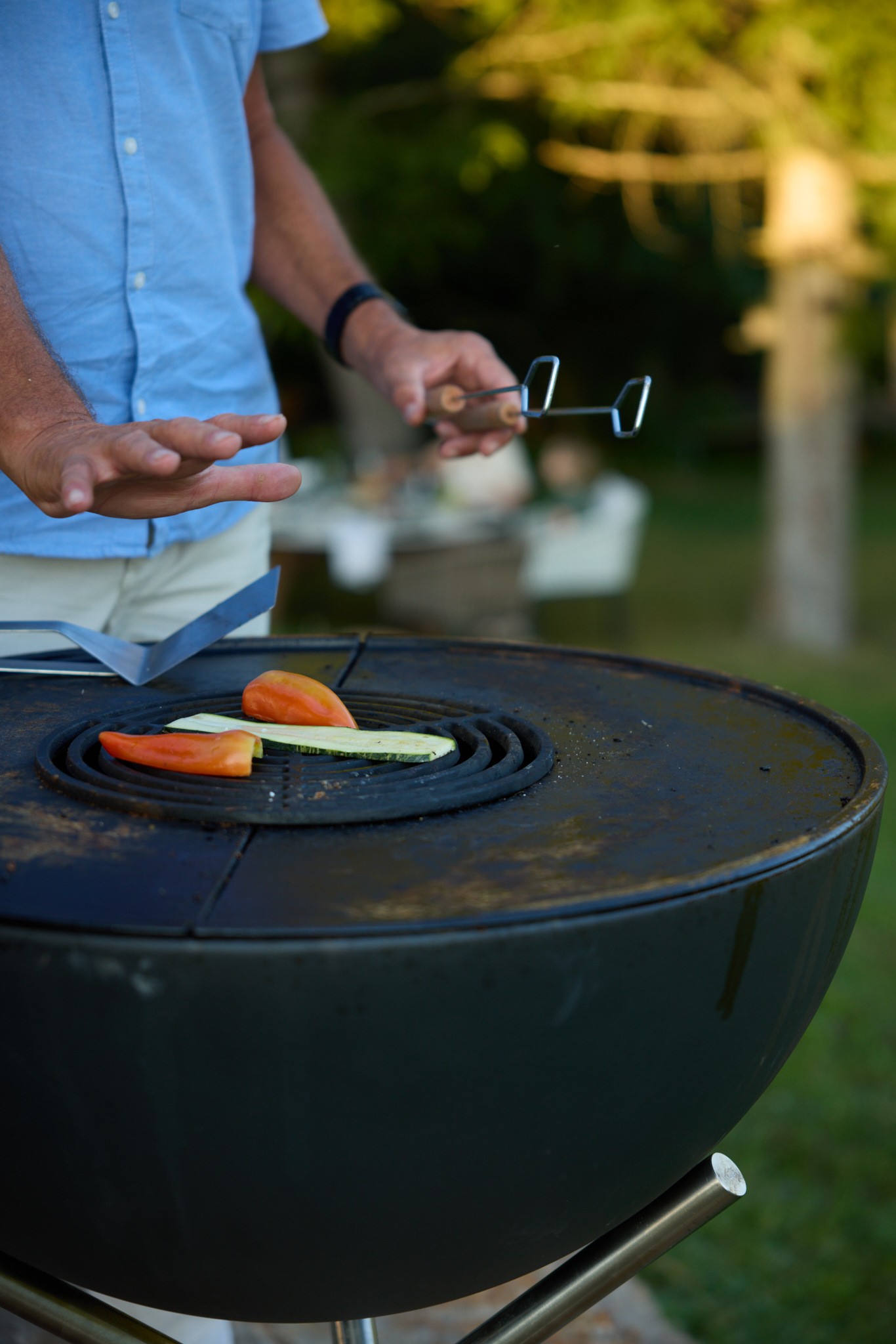 Nahaufnahme von Händen einer Person die alleine vor einem Grill steht und Gemüse wendet.